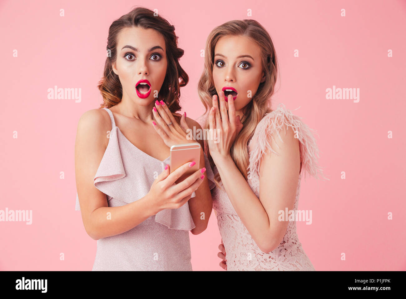 Two shocked elegant women in dresses covering their mouths while using smartphone and looking at the camera over pink background Stock Photo