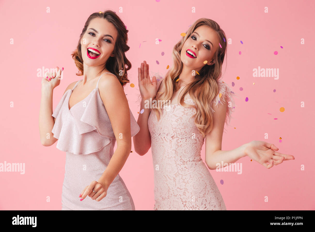 Two cheerful elegant women in dresses having fun together and looking at the camera over pink background Stock Photo