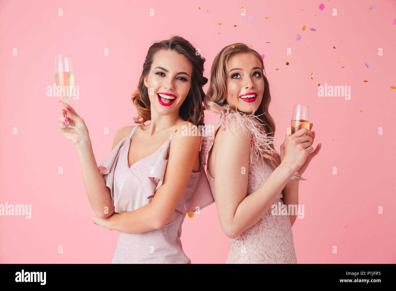 Two happy elegant women in dresses posing together with champagne and looking at the camera over pink background Stock Photo