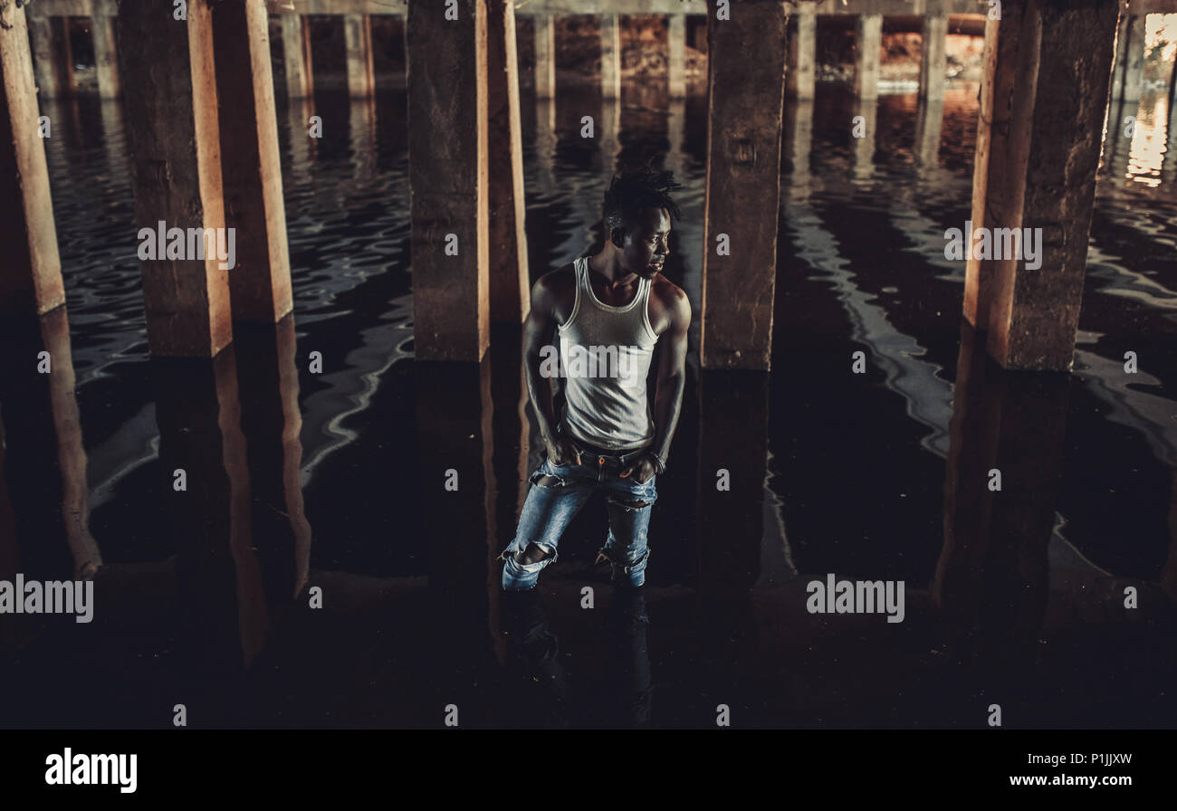 Young African man in white tank top and jeans stands in water under bridge on background of concrete supports. Stock Photo
