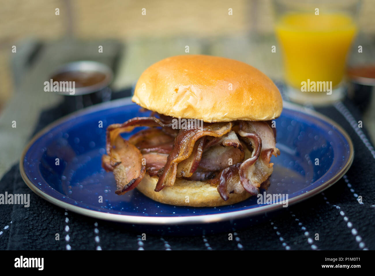 Streaky Bacon in a brioche bun, with ketchup and orange juice in the background. Stock Photo