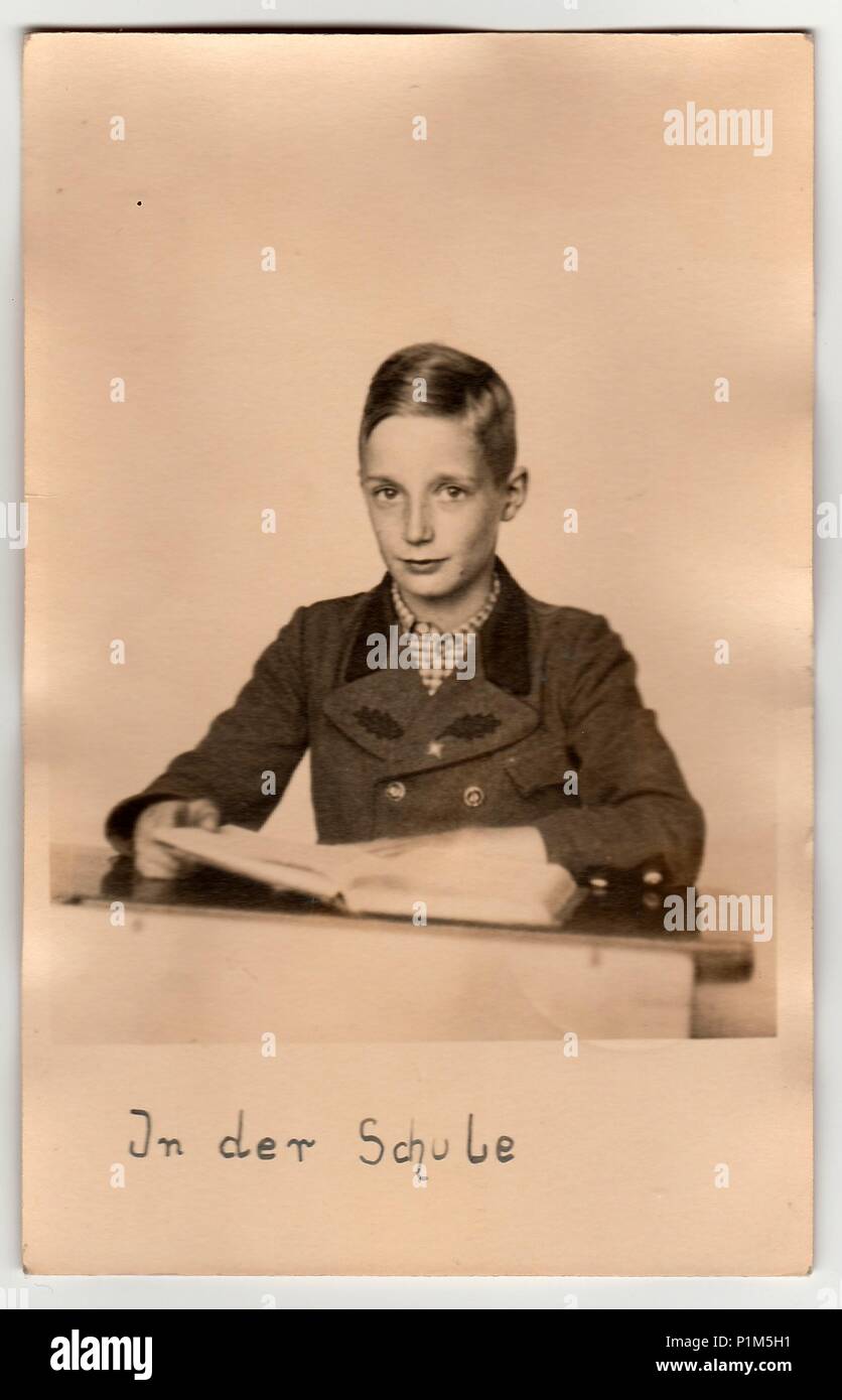 GERMANY - AUGUST 6, 1942: Vintage photo shows young boy (pupil) at the school desk. Pupil wears a period jacket (oaken epaulettes/marks on jacket). On photography is text in German - 'In the school' (translation in English). Stock Photo