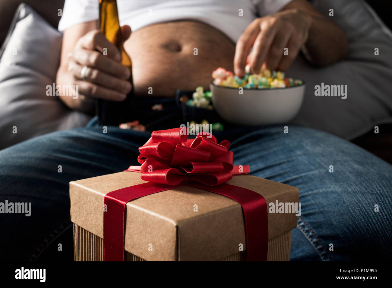 closeup of a gift and a caucasian man with a beer belly, sitting on the sofa, drinking beer and eating popcorn while is watching the television Stock Photo