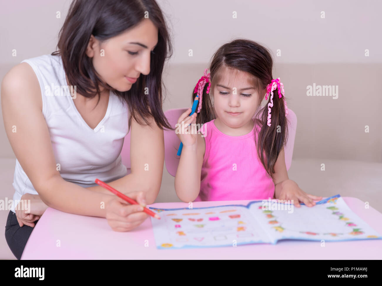 Mother and daughter study activity book with pencils on paper while sitting at pink table.Preschool and kindergarten education at home.Selective focus Stock Photo