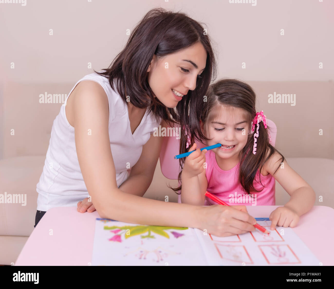 Mother and daughter study activity book with pencils on paper while sitting at pink table.Preschool and kindergarten education at home.Selective focus Stock Photo