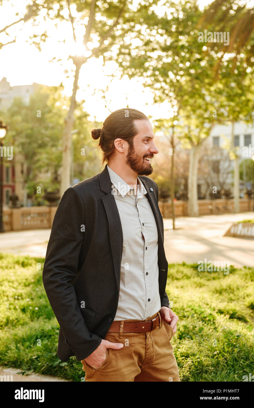 Profile picture of cheerful handsome man with tied hair smiling and standing with hands in pockets in green park during sunny day Stock Photo