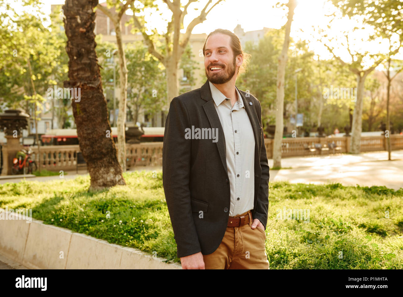 Portrait of positive handsome man 30s with tied hair smiling and standing with hand in pocket in green park during sunny day Stock Photo