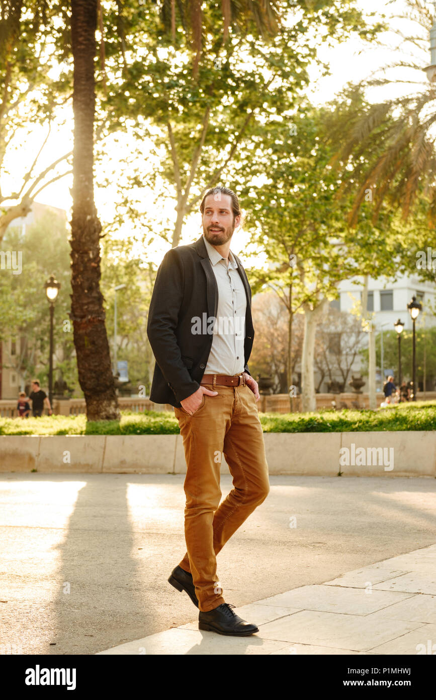 Full length image of unshaved attractive guy 30s with tied hair posing with hands in pockets during walking in green park Stock Photo