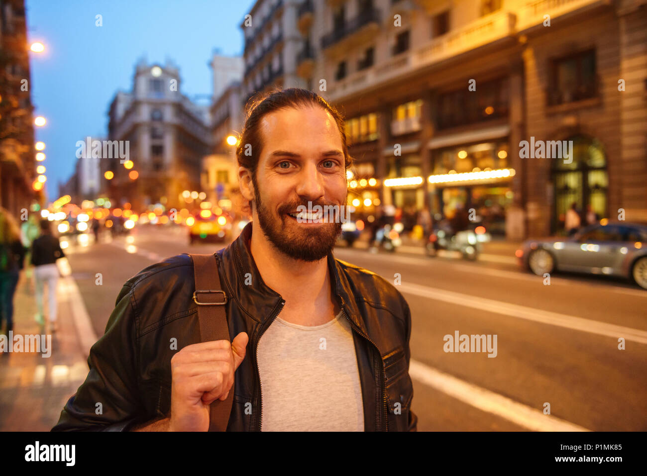 Portrait of joyful handsome man with tied hair and beard smiling while strolling in evening city across main avenue Stock Photo