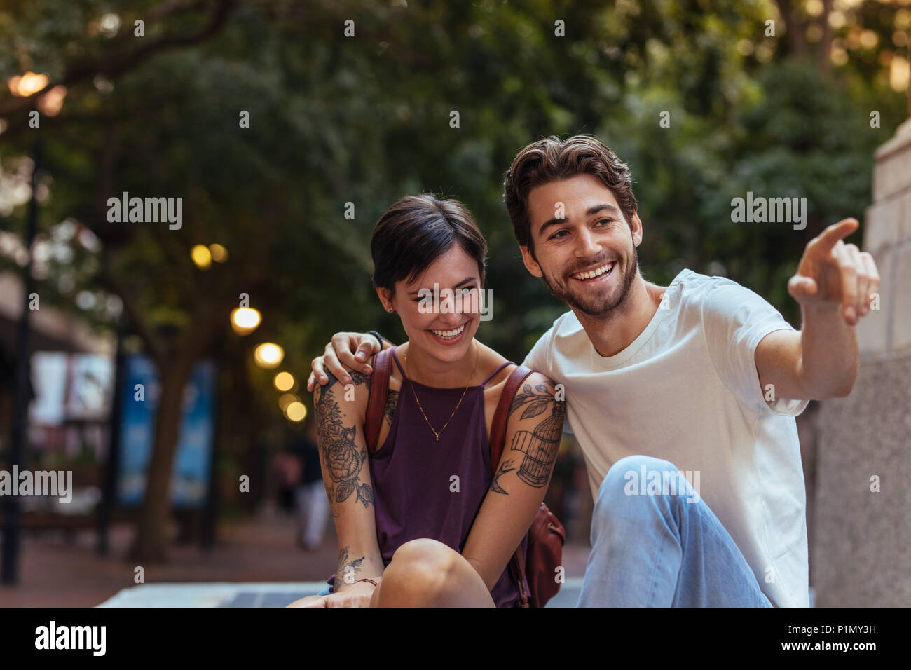 Happy explorer couple sitting outdoors and talking. Man talking to woman with his arm around her shoulders and pointing towards an object. Stock Photo