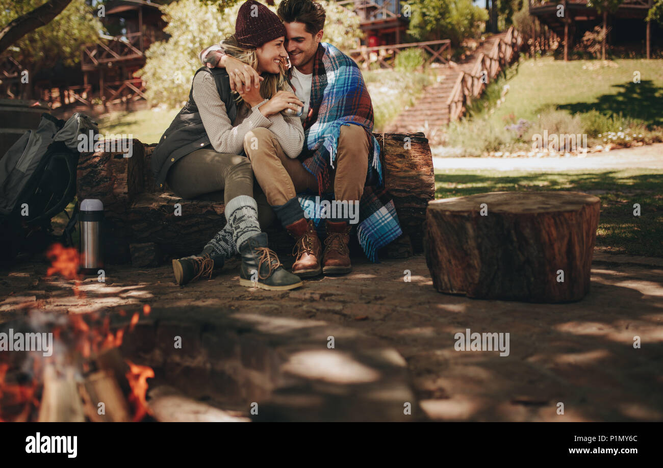 Romantic couple sitting at the campsite. Man and woman sitting on a log at campsite with bonfire in front. Stock Photo