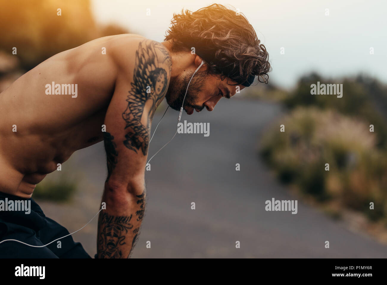 Side view of muscular man taking rest after a run outdoors. Sportsman looking tired after running training on mountain road. Stock Photo