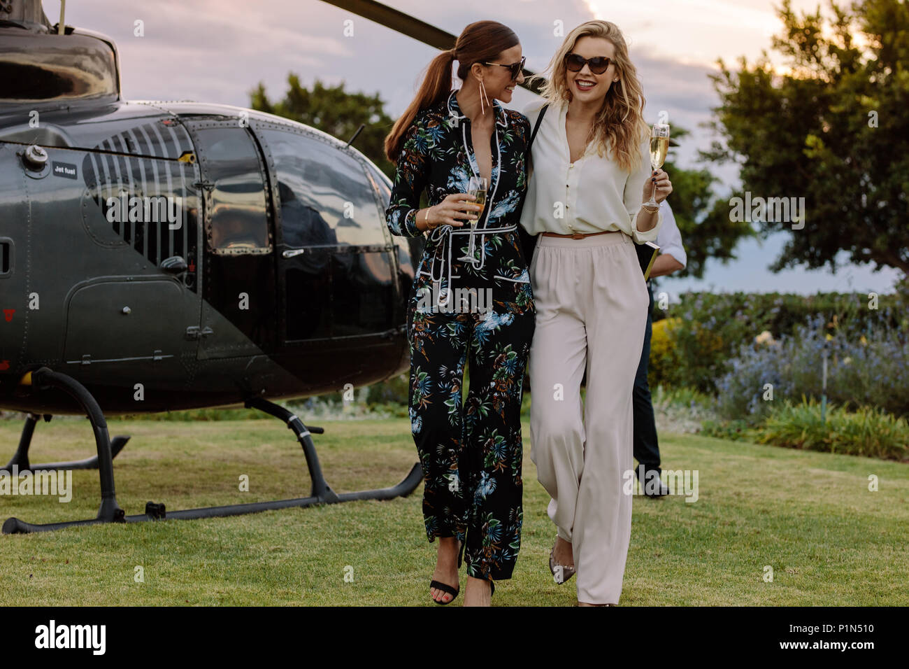 Two young women walking away from a helicopter with a glass of wine in their hands. Best friends walking together with wine and having fun. Stock Photo