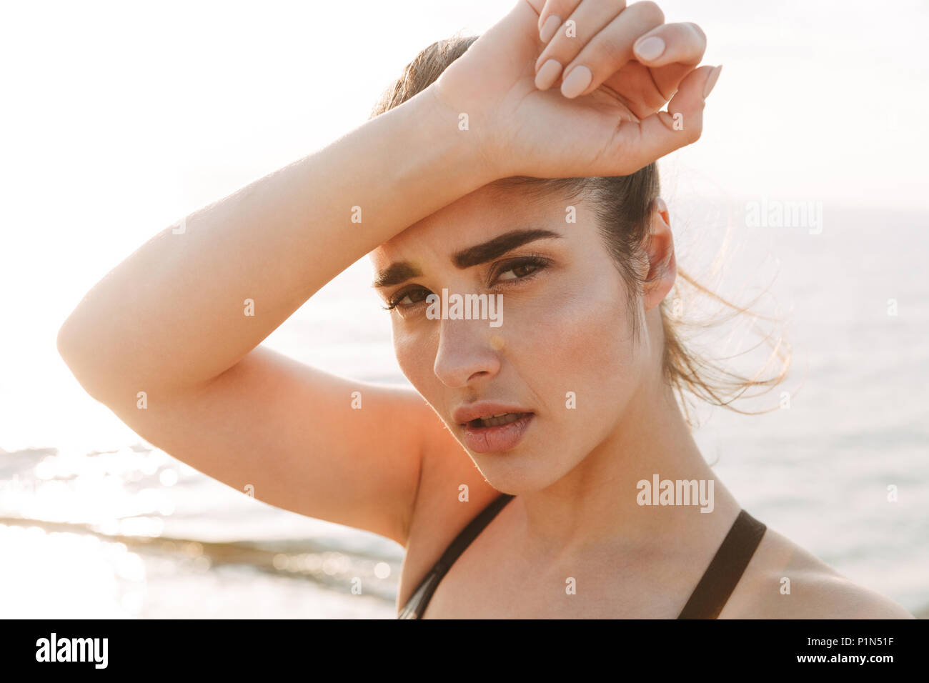 Close up portrait of a tired young sportswoman wiping forehead with her hand at the beach Stock Photo