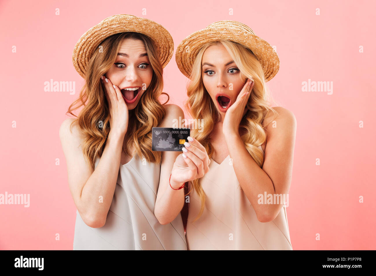 Portrait of two excited young women in summer clothes looking at plastic credit card isolated over pink background Stock Photo