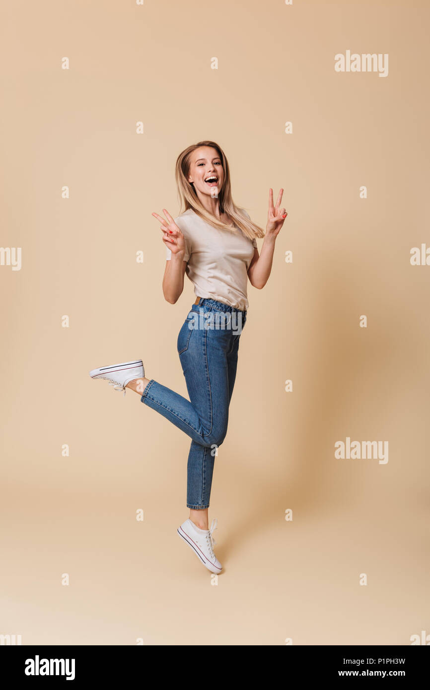Full length image of content easygoing blond woman 20s wearing casual clothing lifting one leg and showing peace sign with two hands isolated over bei Stock Photo