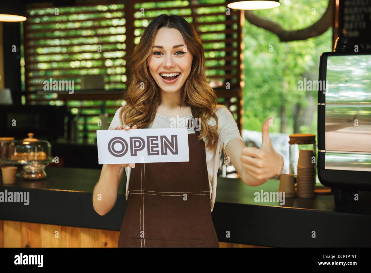 Portrait of a smiling young barista girl in apron holding open sign board while standing at the cafe and showing thumbs up Stock Photo