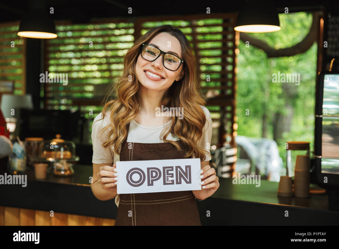 Portrait of a pretty young barista girl in apron holding open sign board while standing at the cafe Stock Photo