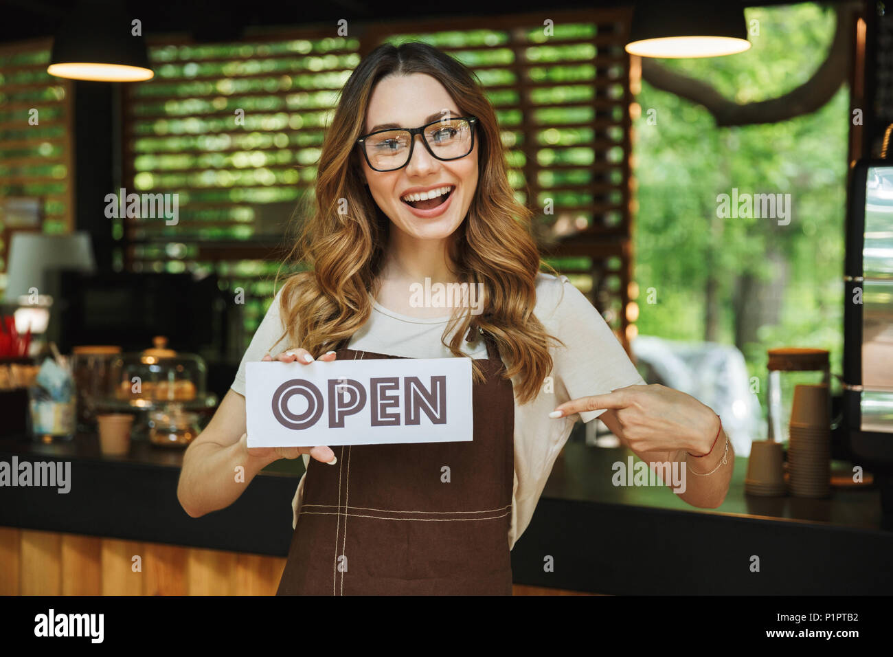 Portrait of a smiling young barista girl in apron holding open sign board while standing at the cafe and pointing Stock Photo
