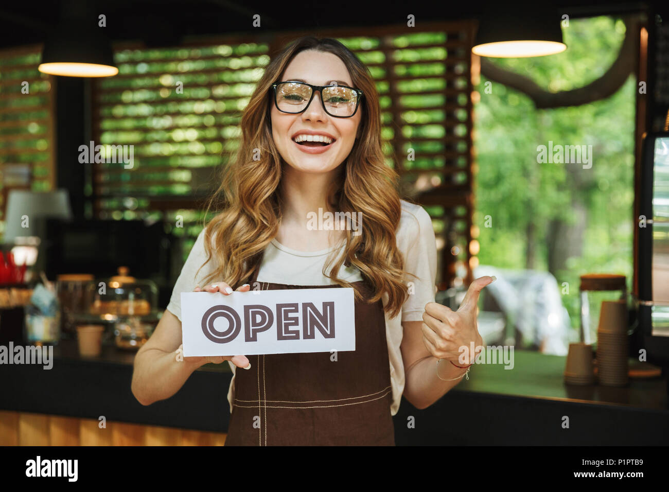 Portrait of a smiling young barista girl in apron holding open sign board while standing at the cafe and showing thumbs up Stock Photo