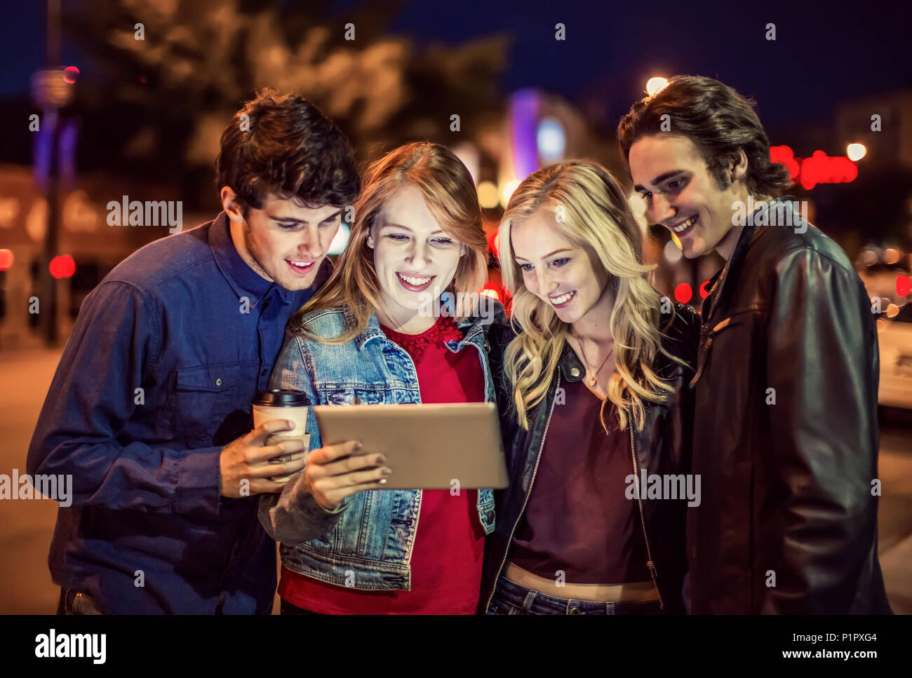 A group of four friends huddle together on a sidewalk looking at a tablet as the glow from the screen lights up their faces; Edmonton, Alberta, Canada Stock Photo