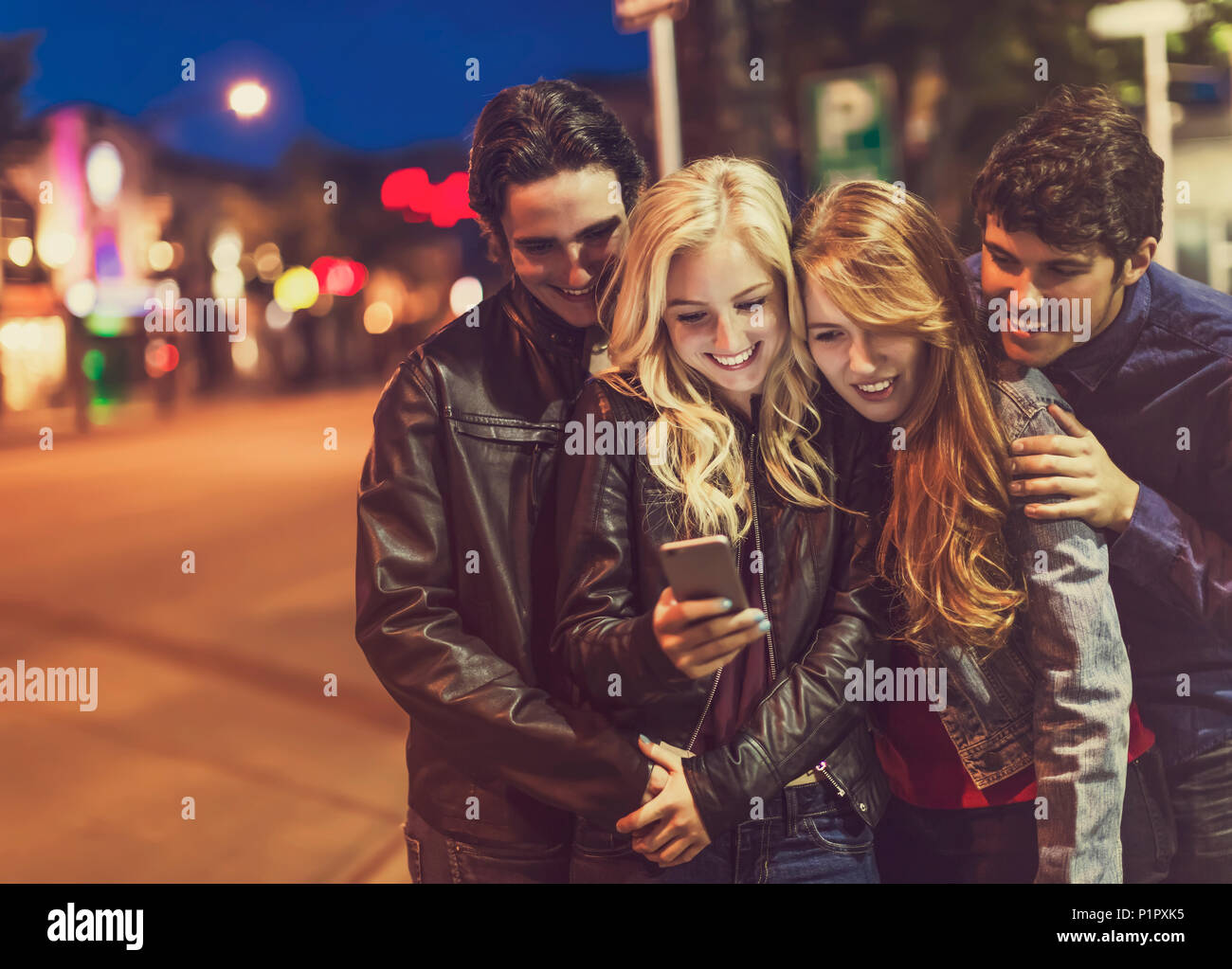 A group of four friends huddle together on a sidewalk looking at a smart phone as the glow from the screen lights up their faces Stock Photo