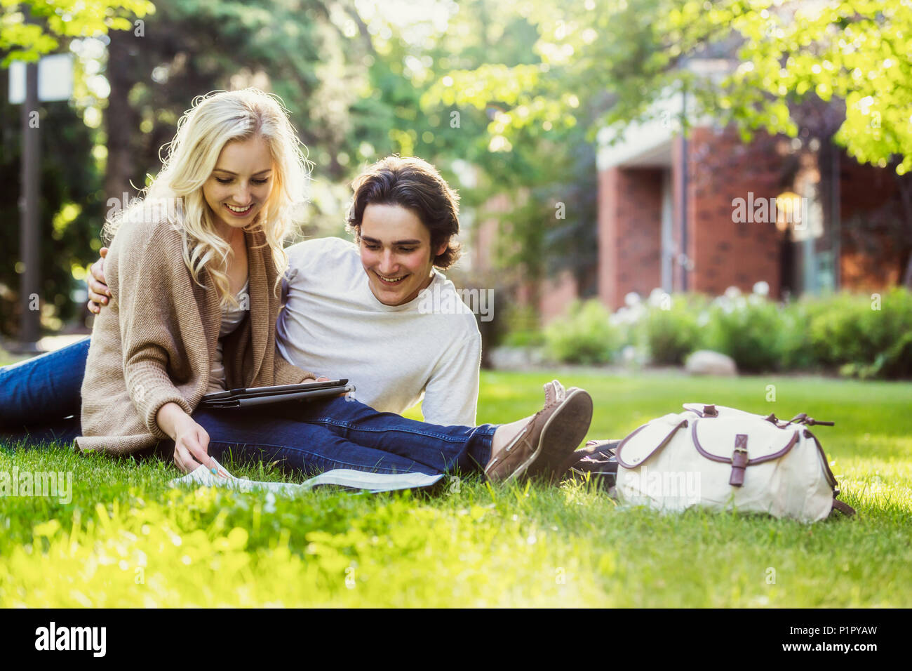 A young couple studying outdoors on the grass on the university campus and looking through a textbook while using a tablet; Edmonton, Alberta, Canada Stock Photo