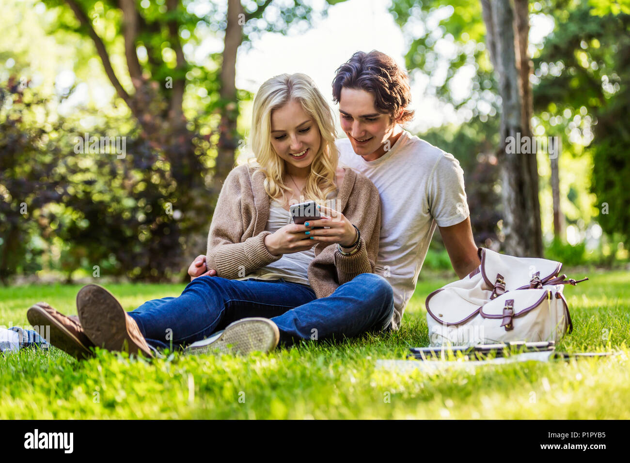 A young couple sits studying on the grass on the university campus with a textbook and tablet while using a smart phone; Edmonton, Alberta, Canada Stock Photo