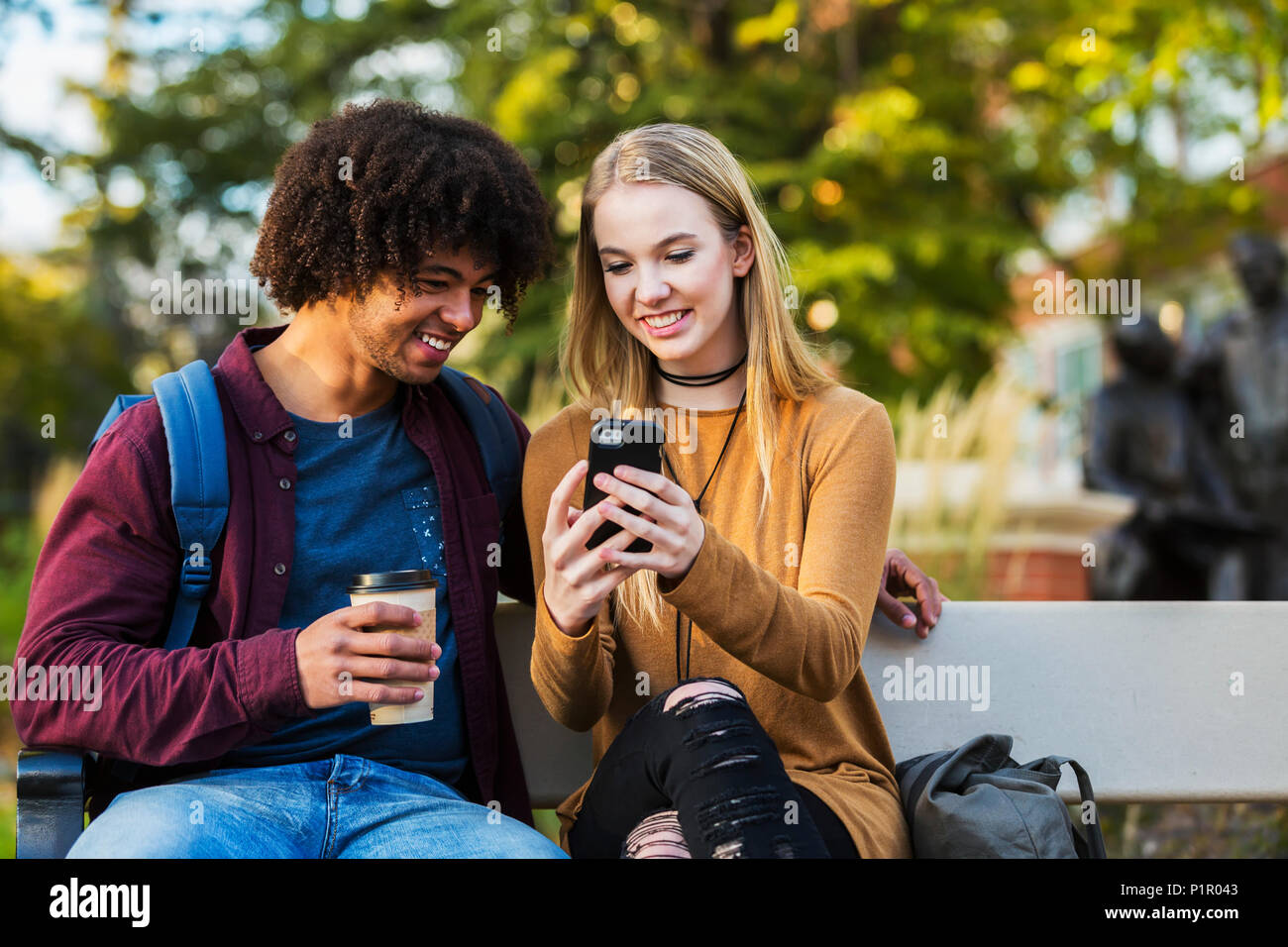 Young dating couple who are university students sitting on a bench on campus and looking at social media on a smart phone; Edmonton, Alberta, Canada Stock Photo