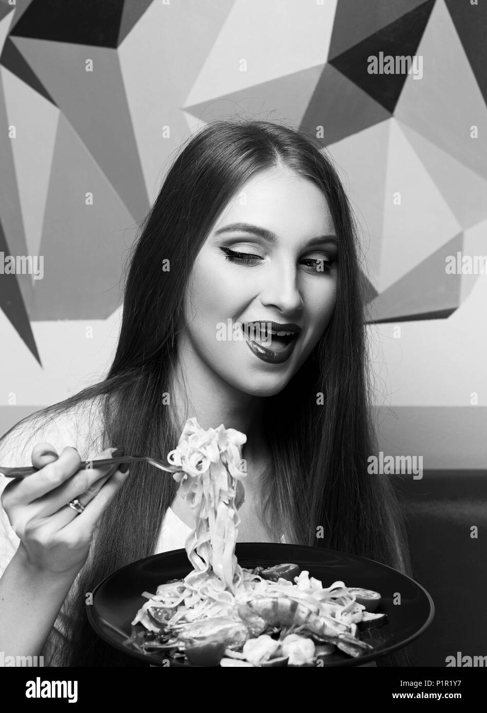 Laughing beautiful long haired woman with expressively opened mouth eating Italian pasta with shrimps and vegetables. Joyful funny young female model  Stock Photo