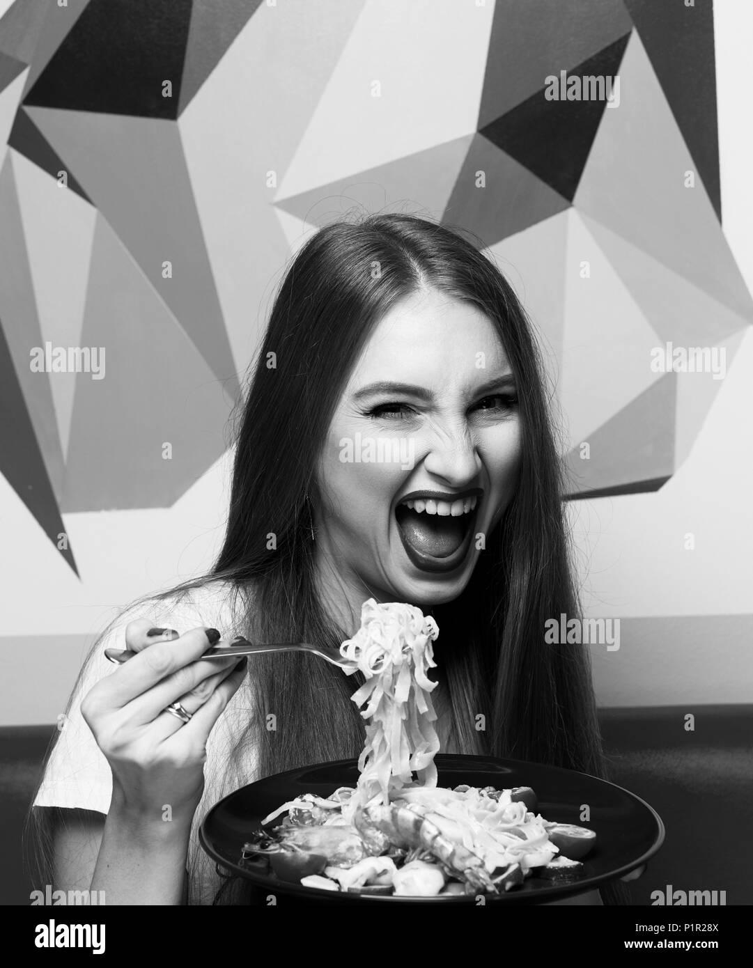 Laughing beautiful long haired woman with expressively opened mouth eating Italian pasta with shrimps and vegetables. Joyful funny young female model  Stock Photo
