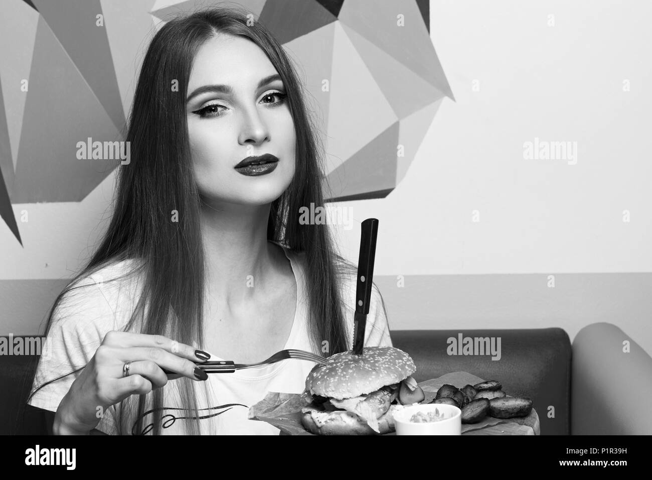 Happy attractive woman holding fork and round wooden plate with appetizing burger and fried potato. Beautiful joyful female model eating cheeseburger. Stock Photo