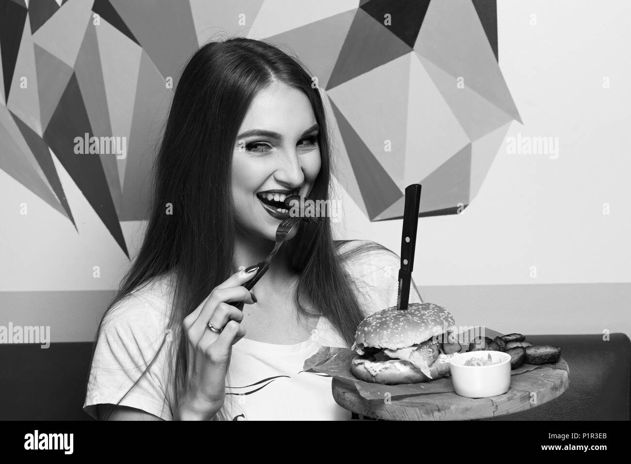 Happy attractive woman holding fork and round wooden plate with appetizing burger and fried potato. Beautiful joyful female model eating cheeseburger. Stock Photo