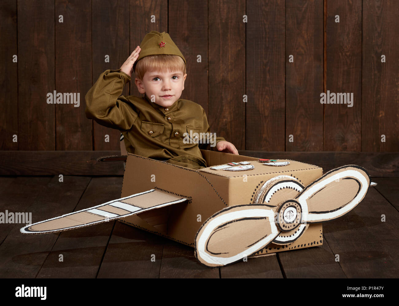 children boy are dressed as soldier in retro military uniforms sit in an airplane made of cardboard box and dreams of becoming a pilot, dark wood back Stock Photo