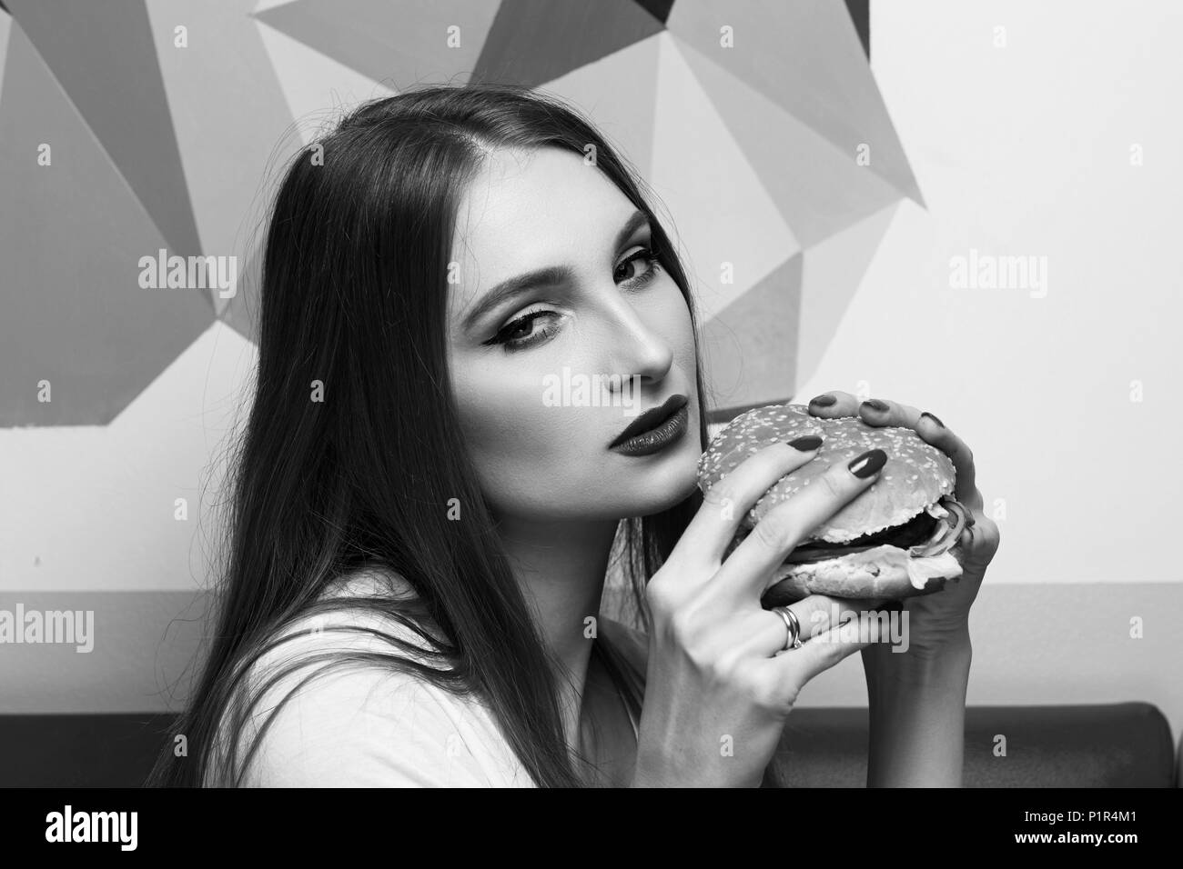 Portrait of gorgeous young woman with closed eyes and red lips holding tasty cheeseburger and biting it. Attractive female model eating burger with pl Stock Photo
