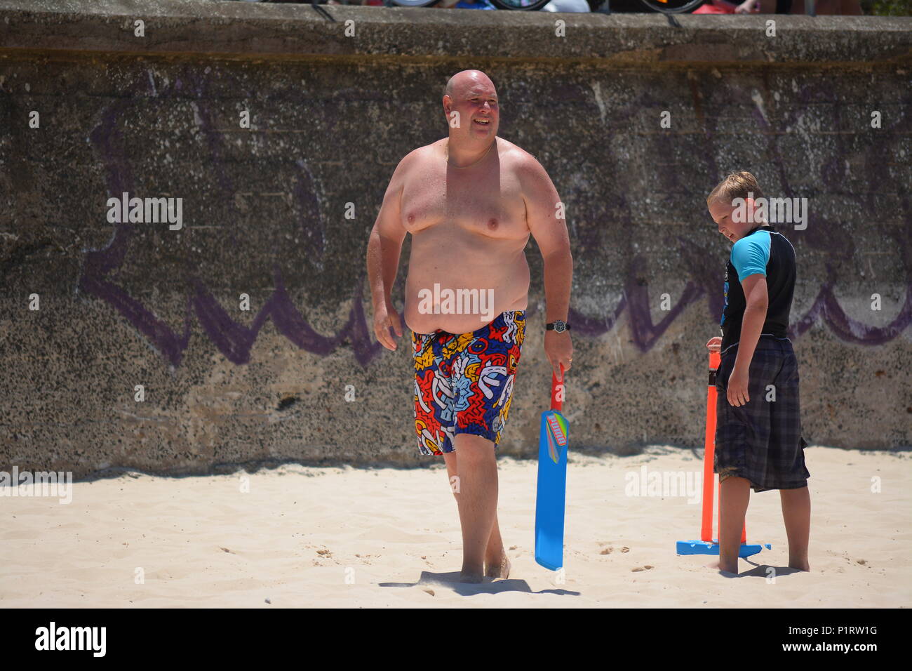 An overweight, obese man playing cricket on beach Stock Photo