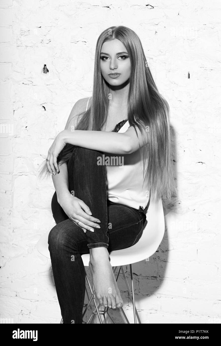 Young beautiful woman with long straight blonde hair in blue jeans and bright blouse sitting on white high bar chair with crossed legs and looking in  Stock Photo