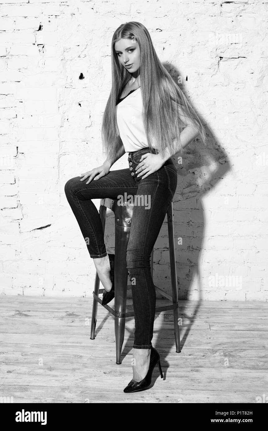 Young beautiful woman with long straight blonde hair in blue jeans and bright blouse sitting on white high bar chair with crossed legs and looking in  Stock Photo