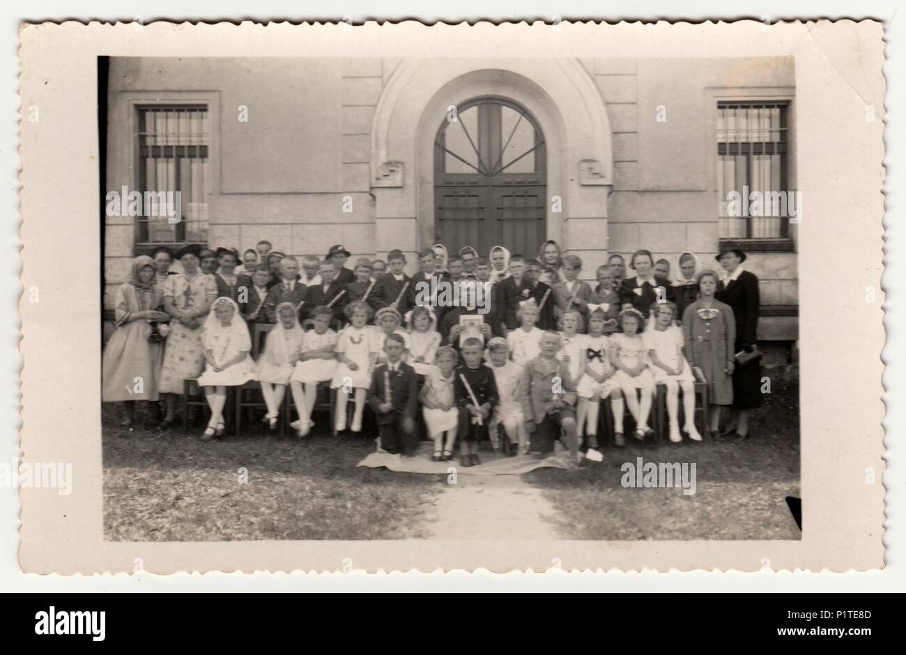 GERMANY - CIRCA 1930s: Vintage photo shows children and their relatives poses after a First Communion in front of the school. Retro black & white photography. Stock Photo