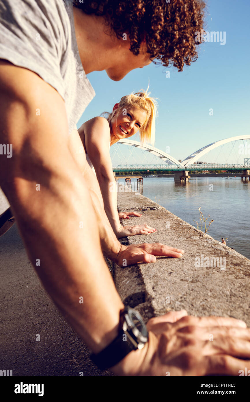 Young happy couple runners training outdoors doing push-ups during outdoor cross training by the river. Stock Photo