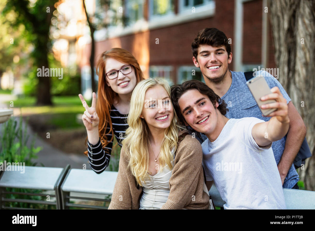 A group of four friends sitting on a bench taking a self-portrait with a smart phone on a university campus; Edmonton, Alberta, Canada Stock Photo