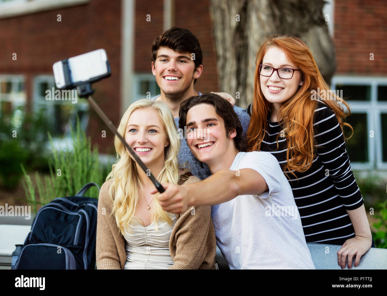 A group of four friends sitting on a bench taking a self-portrait with a smart phone using a selfie stick on a university campus Stock Photo