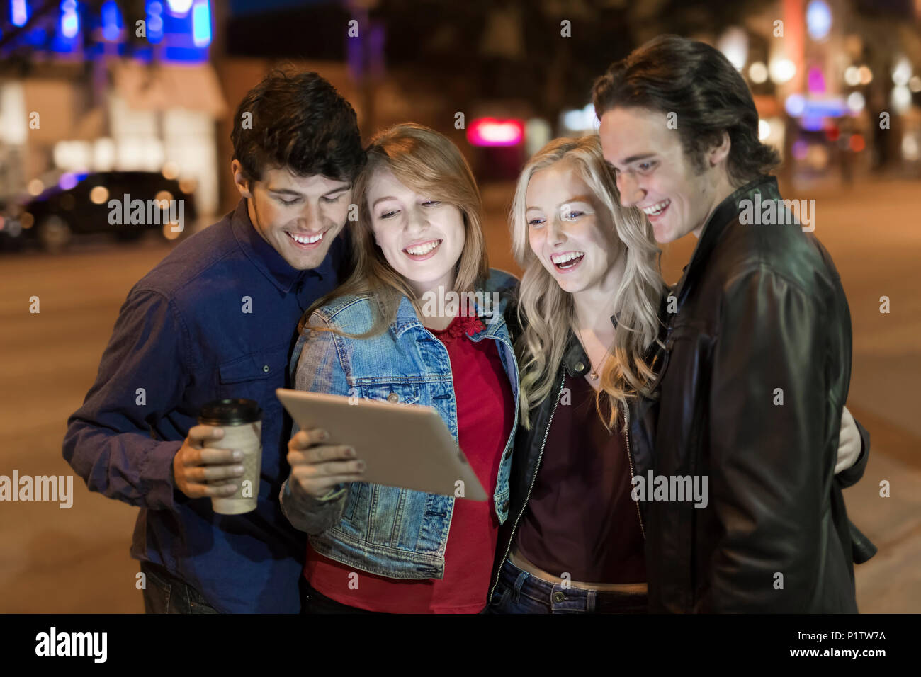 A group of four friends huddle together on a sidewalk looking at a tablet as the glow from the screen lights up their faces; Edmonton, Alberta, Canada Stock Photo