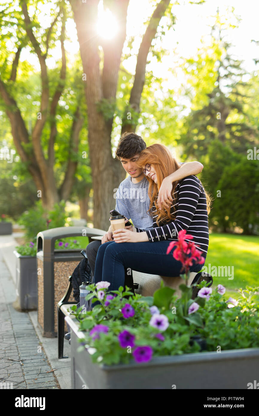 A young couple sitting together on a bench in a park and checking social media on a smart phone; Edmonton, Alberta, Canada Stock Photo