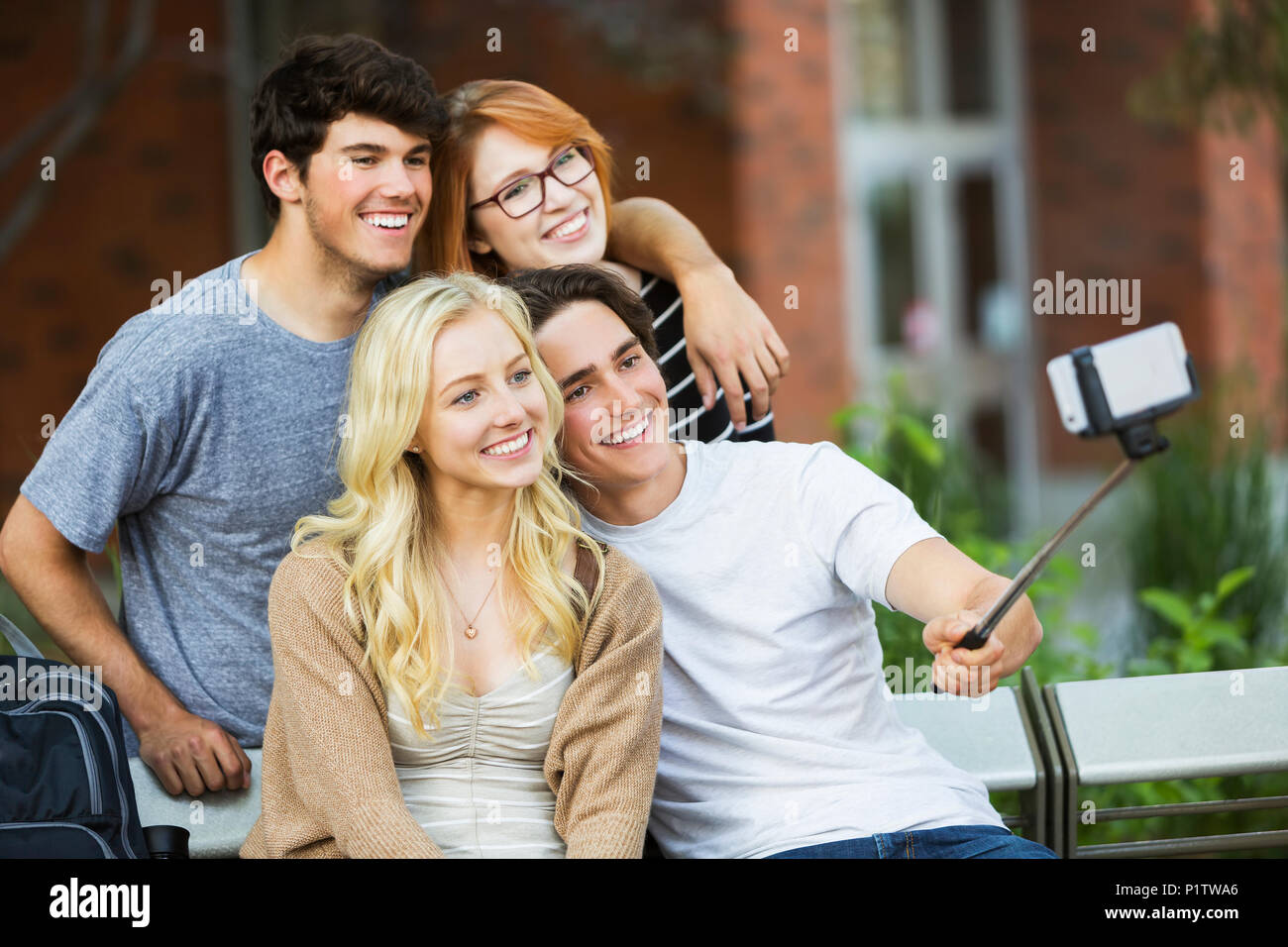 A group of four friends sitting on a bench taking a self-portrait with a smart phone using a selfie stick on a university campus Stock Photo