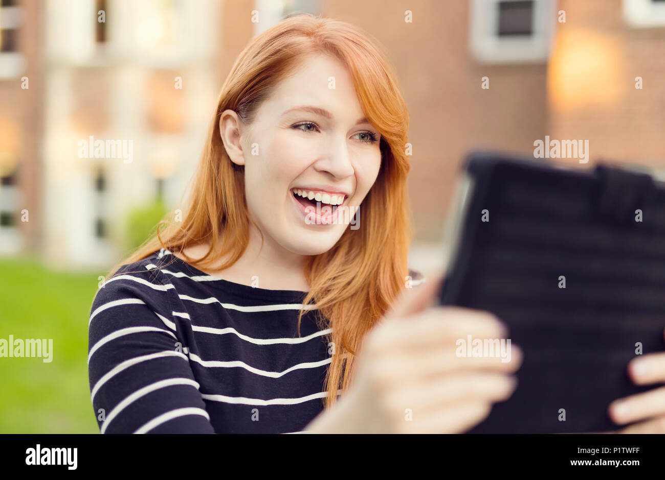 A young woman with red hair smiles big while looking at a tablet; Edmonton, Alberta, Canada Stock Photo