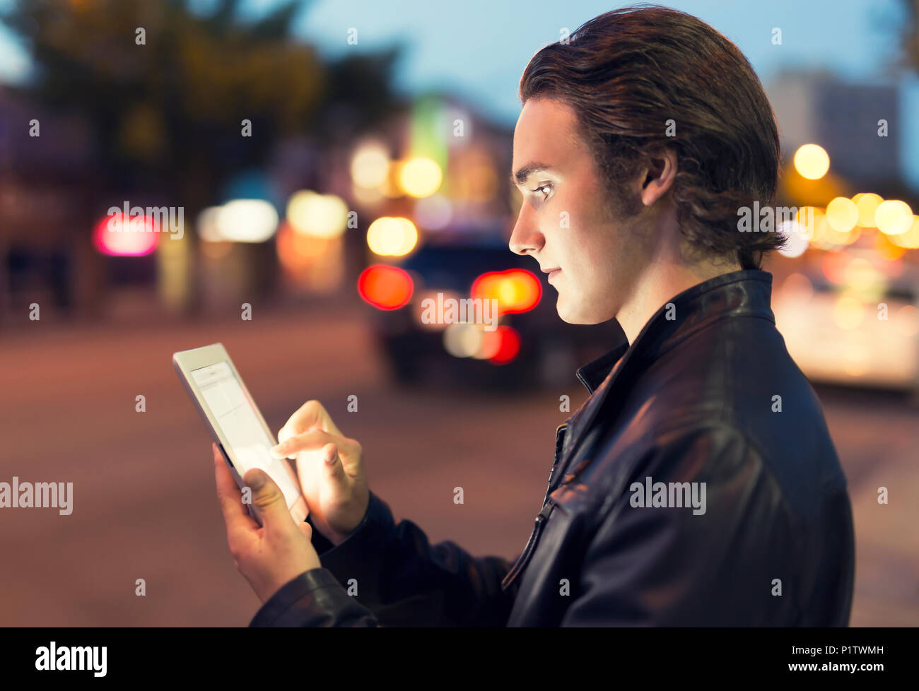 A young man uses a tablet along a street at dusk with the glowing screen illuminating his face; Edmonton, Alberta, Canada Stock Photo