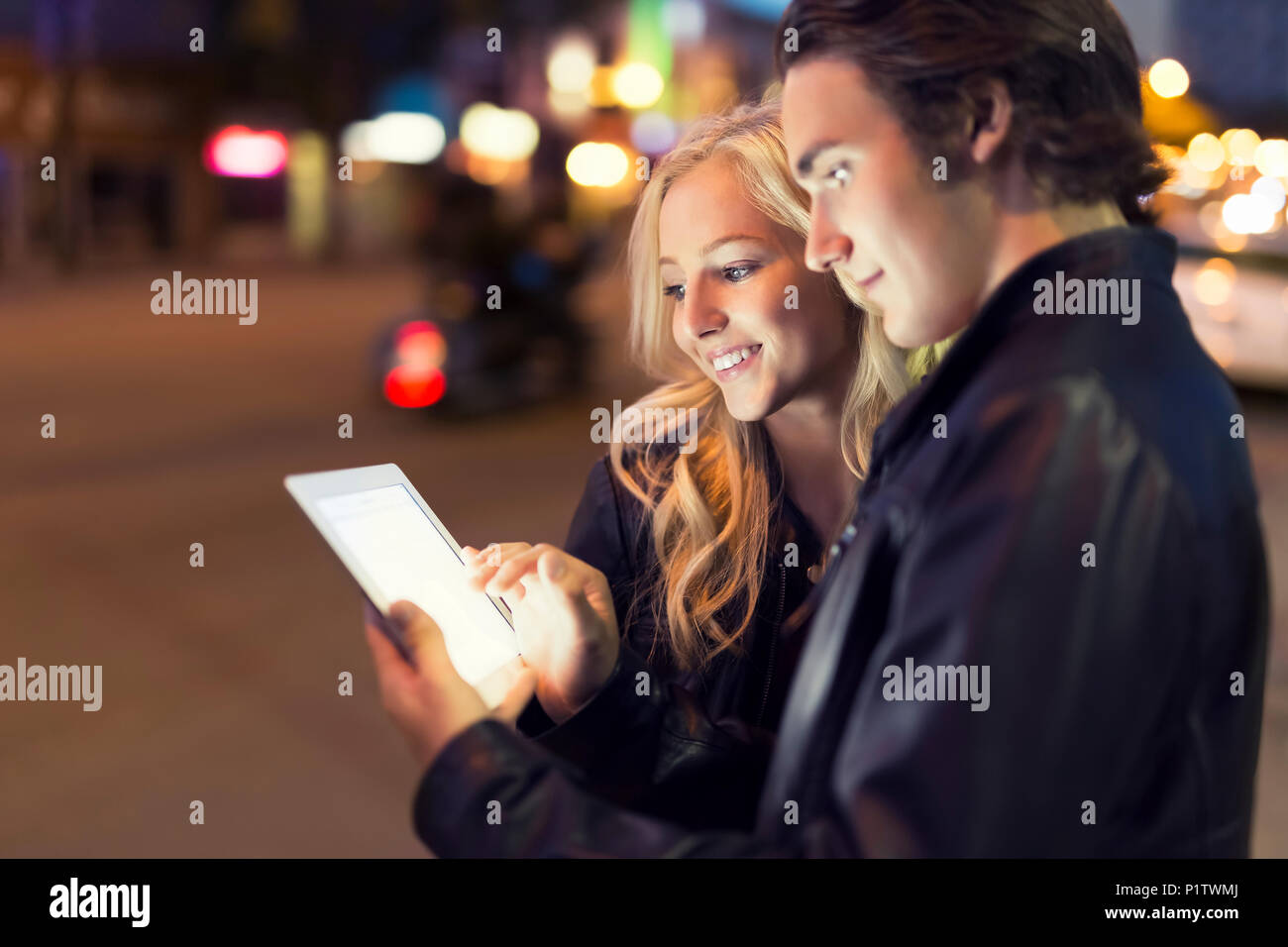 A young couple use a tablet along a street at dusk with the glowing screen illuminating their faces; Edmonton, Alberta, Canada Stock Photo