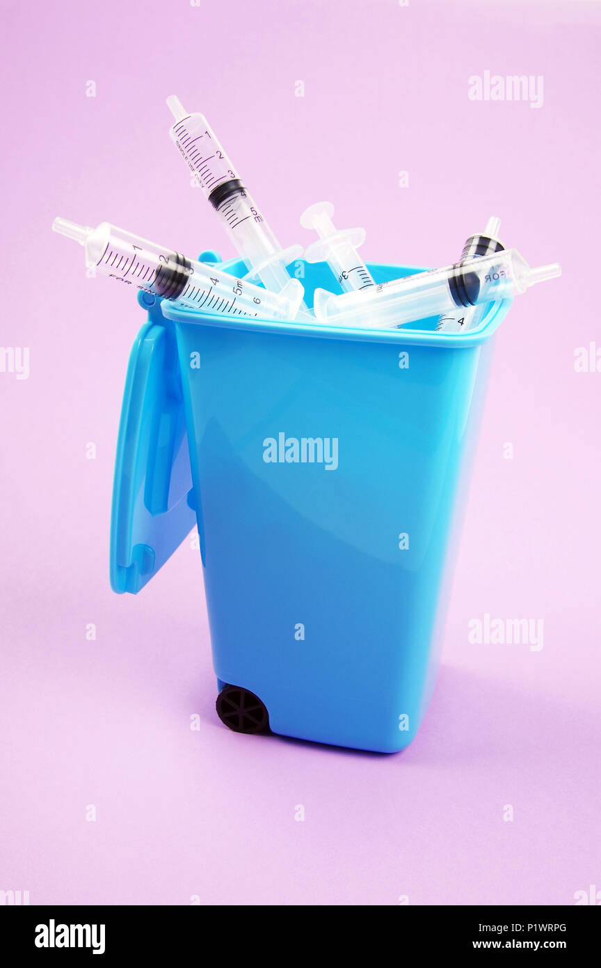 clear plastic medical syringes inside a blue rubbish bin with open lid on a pink tint background Stock Photo