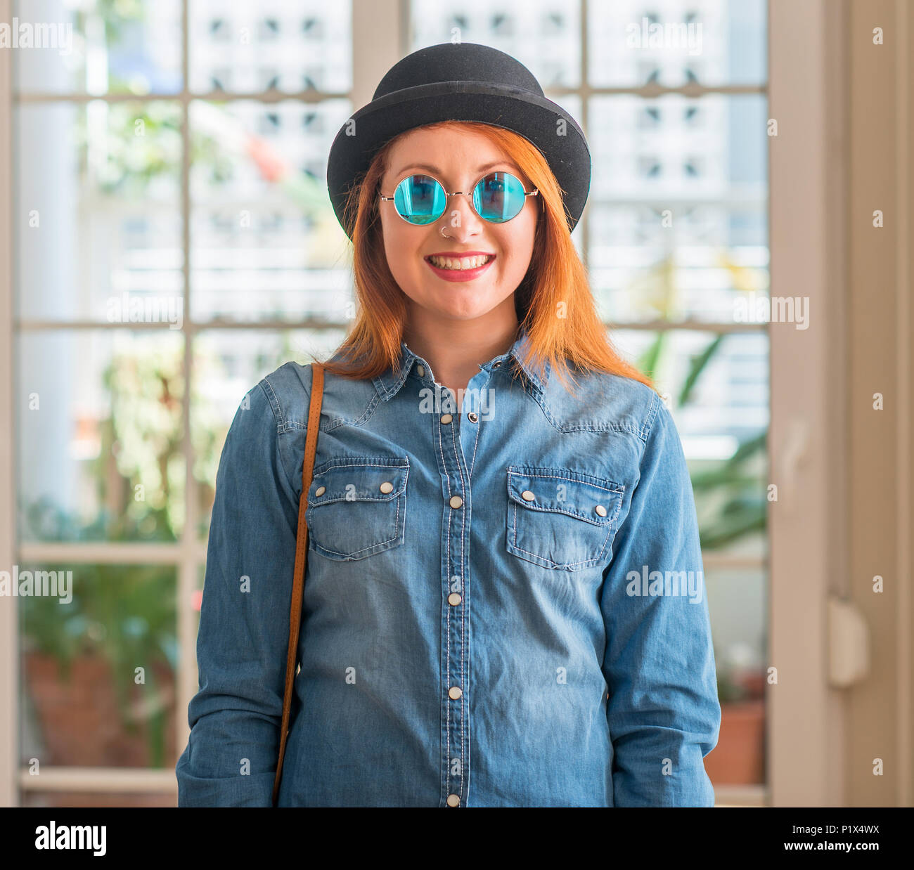 Stylish redhead woman wearing bowler hat and sunglasses with a happy and cool smile on face. Lucky person. Stock Photo